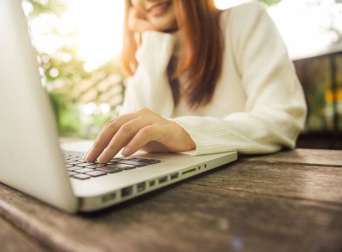 Young woman with light brown hair is pictured from chin down working at a laptop as she applies to college.