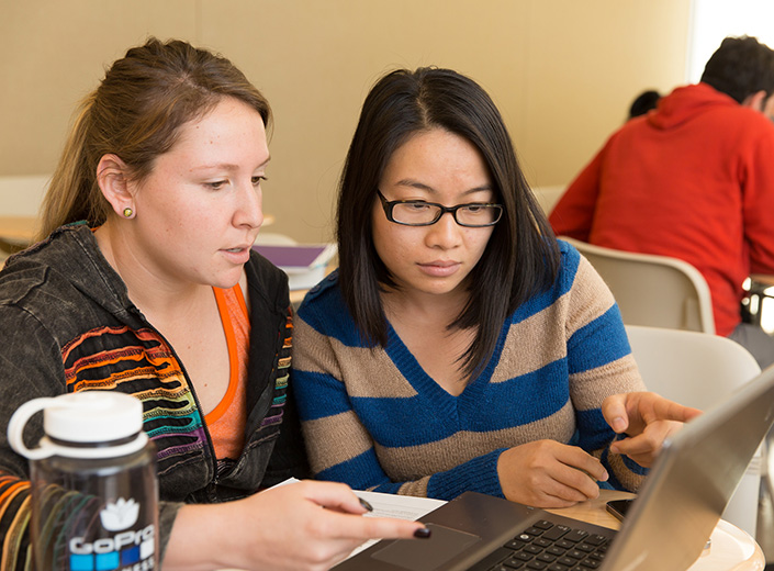 Two young women work together at a computer. One is white with light brown hair in a ponytail. The other is of Asian descent with black plastic glasses. She points with a pencil toward the screen.