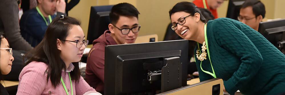 Students work in a computer lab. A female instructor with dark hair in a ponytail leans around the desk to demonstrate something to a female student in glasses.