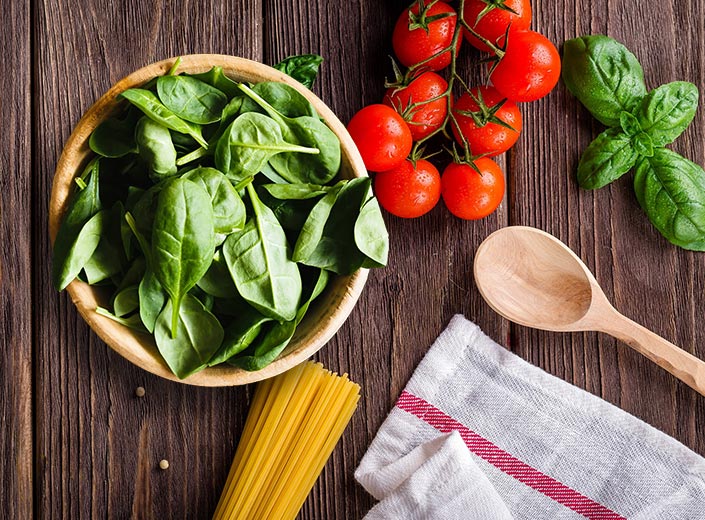 Spinach, tomatoes, spaghetti noodles (dry), and basil leaves are laid out on a dark wooden table top.