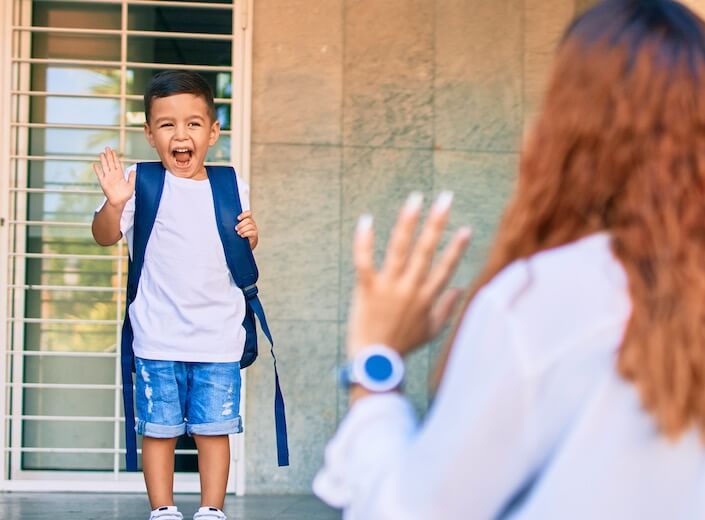 Latino toddler waves goodbye from steps of school to mom. 
