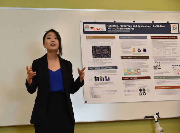A Chemistry student, female of Asian descent, wears a black suit and presents her project at the front of a classroom.