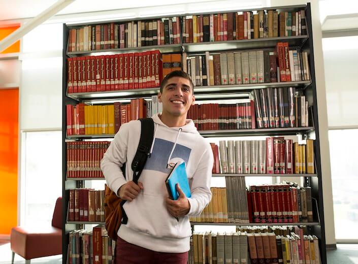 Latino college student in white hoodie holds a book and has backpack slung over his shoulder. He is standing in front of library stacks.