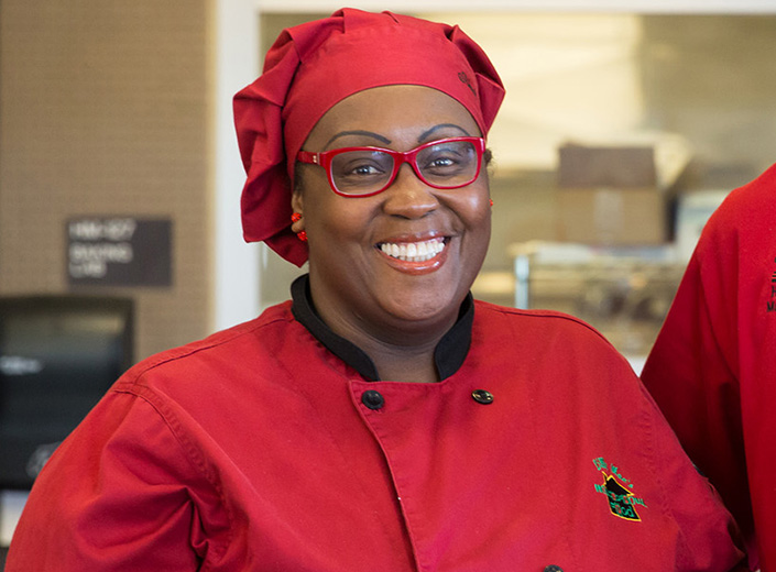 African-American woman chef wear red uniform and black glasses. She stads in a kitchen.