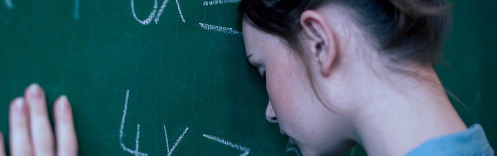 A young woman with light skin and brown hair pulled back in a bun leans against a blackboard (forehead is touching it) that has equations written on it.