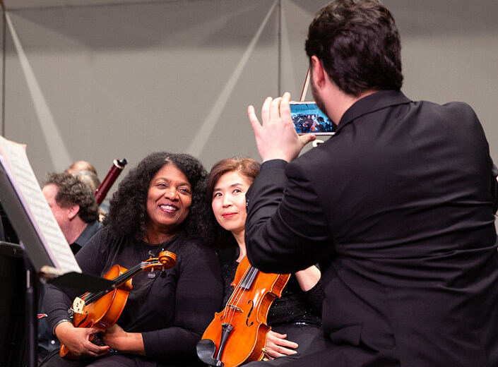 Two women pose together at a Mission Symphony orchestra in black clothes holding their violins.