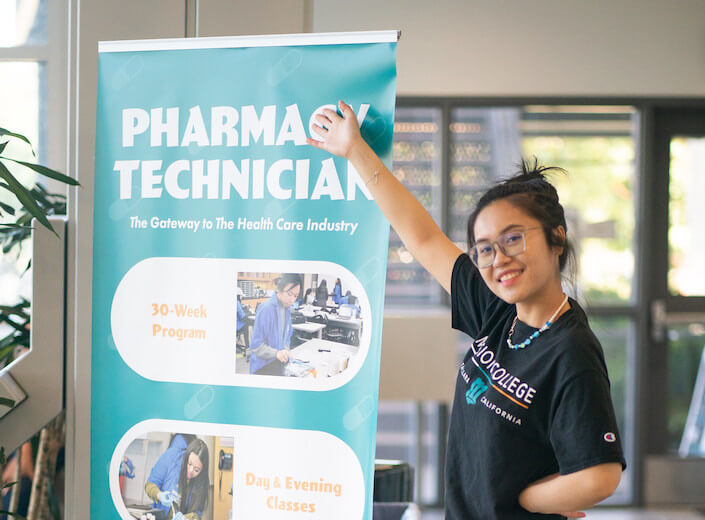 Young woman of Asian descent wears a black tshirt with "Mission College" written in teal. She gestures to a tall sign that reads ha"Pharmacy Technician".
