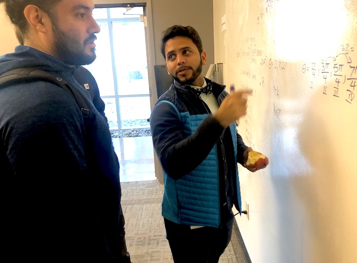 A male college student of South-Asian descent performs some math equations on a whiteboard.