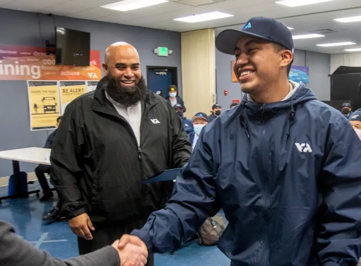 VTA Apprenticeship grad in blue windbreaker and baseball hat shakes hand of female instructor in a classroom. He is smiling.