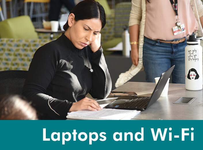 A woman is pictured working at a laptop in the library. She is looking at her notebook. She has black hair and olive skin. Her upper ear is pieced. "Laptops and Wi-Fi" is written in white text across the bottom of the image across a strip of teal.