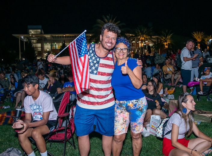 Man in red and white striped shirt with blue shorts holds a flag and poses with his arm around an older lady at an outdoor concert.