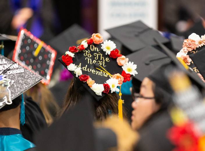 Students are pictured from the back of the room, mostly visible are their decorated commencement caps.
