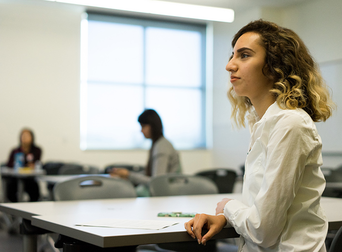Young woman with curly light brown hair, olive skin, and white long-sleeved shirt pays attention to a lesson in a classroom.