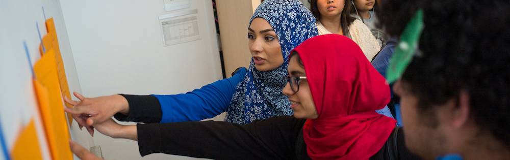 Two young women in colorful hijabs work together at a white marker board.