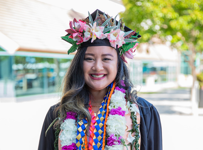 Female graduate of AAPI descent wears her cap and gown. The cap and gown are decorated with lots of flowers.