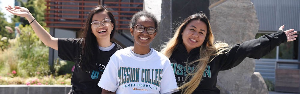 Three young women in Mission College-branded shirts pose outside on campus.