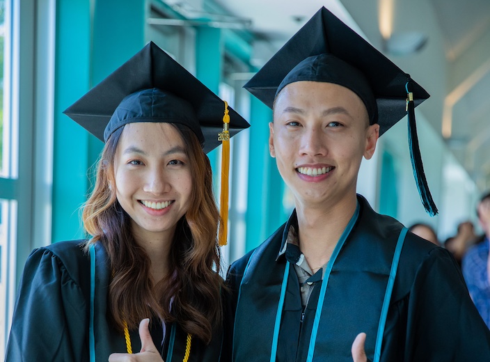Young Asian man and woman smile wearing commencement cap and gown.