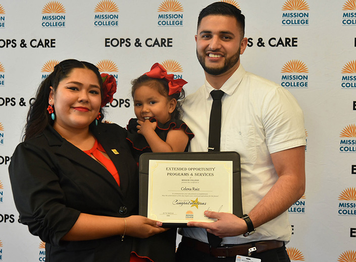 A latinx family (father, mother, and young child) pose in from an EOPS/CARE backdrop at an event.
