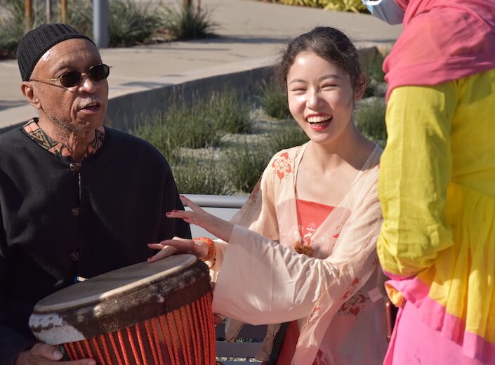 Female student laughs and drums on a djembe held by an instructor in traditional African clothing. They are seated outside at an event on campus for International Day.