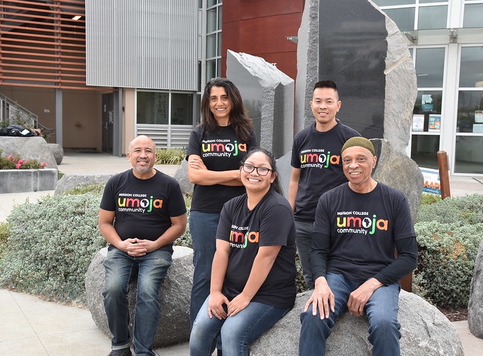 Umoja staff and faculty wear black tshirts with multi-colored letters reading "Umoja" on the front. They sit outside the Student Engagement Center on the MIssion College campus in Santa Clara, California.