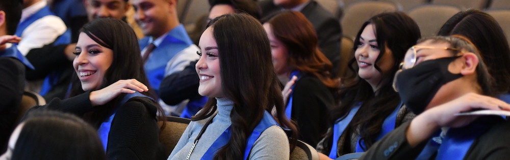 Students in business formal attire and bright blue sashes attend a graduation ceremony in an auditorium for the Year Up program.